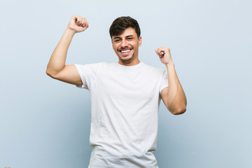 Young caucasian man wearing a white tshirt celebrating a special day, jumps and raise arms with...