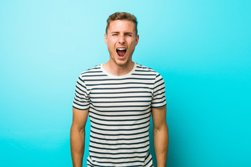Young caucasian man against a blue wall screaming very angry and aggressive.
