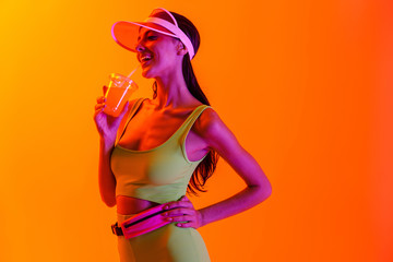 Emotional optimistic stylish girl in sport lifestyle clothes and transparent cap drinking juice cocktail isolated over orange background with neon lights.