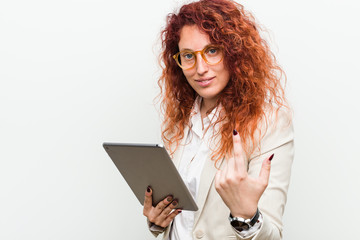 Young caucasian business redhead woman holding a tablet pointing with finger at you as if inviting come closer.