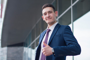 Fototapeta premium Happy confident cheerful businessman in suit and tie showing giving thumb up, like, seal of approval. Portrait of successful guy outdoor office building, business centre. Making like gesture.