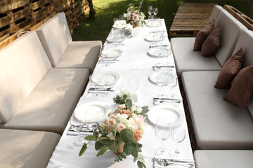 Wedding table setting decorated with fresh flowers in a brass vase. Wedding floristry. Banquet table for guests outdoors with a view of green nature. Bouquet with roses, eustoma and eucalyptus leaves