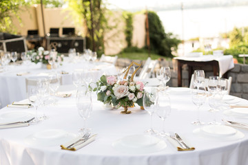 Wedding table setting decorated with fresh flowers in a brass vase. Wedding floristry. Banquet table for guests outdoors with a view of green nature. Bouquet with roses, eustoma and eucalyptus leaves