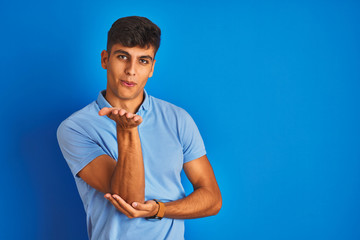 Young indian man wearing casual polo standing over isolated blue background looking at the camera blowing a kiss with hand on air being lovely and sexy. Love expression.