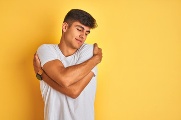 Young indian man wearing white t-shirt standing over isolated yellow background Hugging oneself...