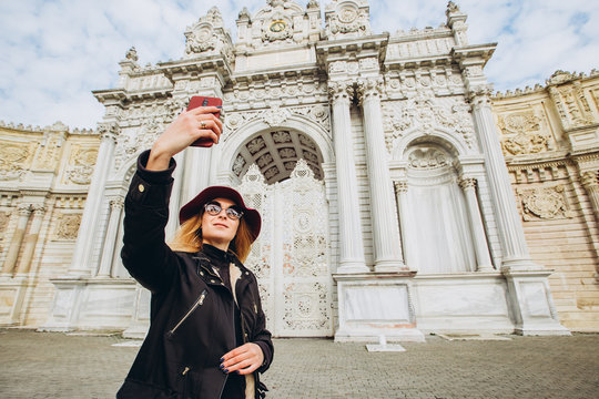 girl takes a selfie in the square in front of Dolmabahce, Istanbul, Turkey. A young girl tourist in a hat and coat is photographed on the phone in front of the Sultan's Palace in Istanbul.