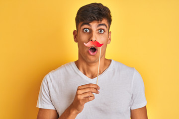 Young indian man holding fanny mustache standing over isolated yellow background scared in shock...