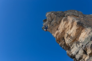 girl climber hanging on the edge of a cliff upside down. Young athletic female rock climber. Mountains and blue sky on the background. Copy space