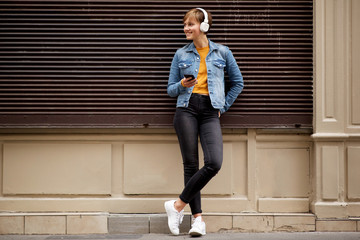 Full body cool young woman standing outside with mobile phone and headphones