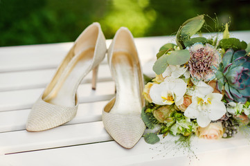 A bouquet of colorful flowers lies on a white bench. Bouquet decorated with green leaves. Exquisite women's high-heeled shoes stand close by.