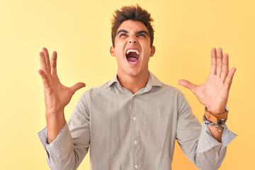 Fototapeta premium Young handsome man wearing casual shirt standing over isolated yellow background crazy and mad shouting and yelling with aggressive expression and arms raised. Frustration concept.