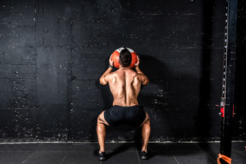 Young strong sweaty focused fit muscular man with big muscles doing throwing medicine ball up on the wall for training hard core workout in the gym real people selective focus