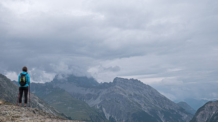 A lone female hiker looks out over some high alpine mountains and valley. Storm clouds gather over the high peaks as she ponders the valley views.