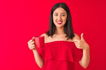 Young beautiful chinese woman drinking cup of coffee standing over isolated red background happy with big smile doing ok sign, thumb up with fingers, excellent sign