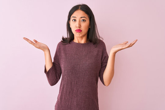Young chinese woman wearing purple sweater standing over isolated pink background clueless and confused expression with arms and hands raised. Doubt concept.
