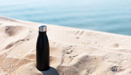 Matte black steel thermo bottle for water in sand of the beach on background of sea. Empty blank...