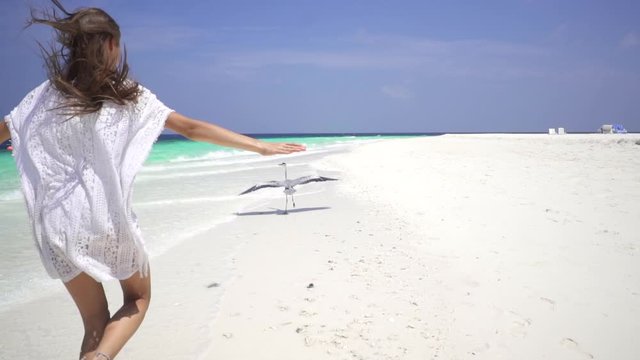 Happy lady in white dress runs after a gray heron along a white sandy beach in the Maldives. A heron flies over the shore. Slow motion. Bora Bora, French Polynesia. Travel motivation.