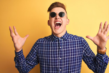 Naklejka premium Young handsome man wearing casual shirt and sunglasses over isolated yellow background crazy and mad shouting and yelling with aggressive expression and arms raised. Frustration concept.