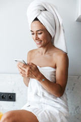 Smiling happy woman in towel after spa procedure shower indoors at home at the kitchen using mobile phone.