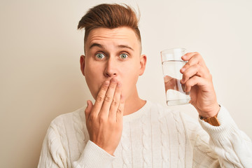 Young handsome man drinking glass of water standing over isolated white background cover mouth with...