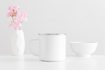Enamel mug mockup with a bouquet of a pink oleander in a vase and a bowl on a white table.
