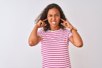 Young brazilian woman wearing pink striped t-shirt standing over isolated white background covering ears with fingers with annoyed expression for the noise of loud music. Deaf concept.