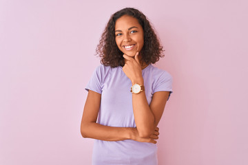 Young brazilian woman wearing t-shirt standing over isolated pink background looking confident at the camera smiling with crossed arms and hand raised on chin. Thinking positive.