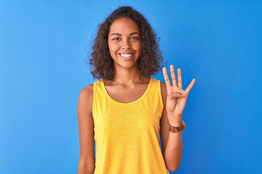 Young brazilian woman wearing yellow t-shirt standing over isolated blue background showing and pointing up with fingers number four while smiling confident and happy.