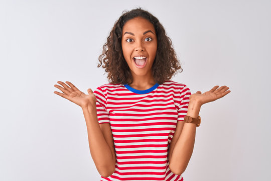 Young brazilian woman wearing red striped t-shirt standing over isolated white background celebrating crazy and amazed for success with arms raised and open eyes screaming excited. Winner concept