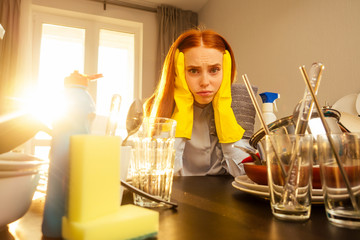 unhappy redhaired ginger beautiful woman wearing yellow glove and washing pile mountains of dirty dishes: cups,pan,saucepan in kitchen