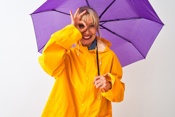 Middle age woman wearing rain coat and purple umbrella over isolated white background with happy face smiling doing ok sign with hand on eye looking through fingers