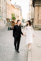 Tender Asian couple in love - bride in white dress and groom in a black suit on their wedding walk in the old city center. Historical ancient buildings on the background