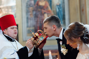 Bride and groom kissing golden crowns from priest hand during wedding ceremony. Spiritual couple. Wedding matrimony in church. Emotional romantic moments