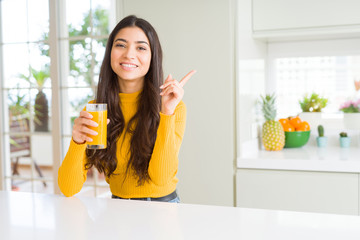 Young woman drinking a glass of fresh orange juice very happy pointing with hand and finger to the...