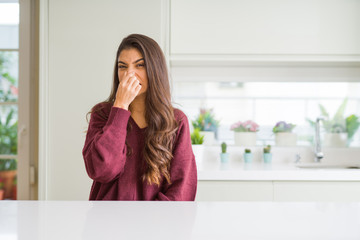 Young beautiful woman at home smelling something stinky and disgusting, intolerable smell, holding breath with fingers on nose. Bad smells concept. - obrazy, fototapety, plakaty