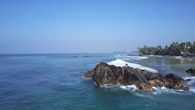 sporty girl stretches in yoga pose standing on grey rocky cliff at ocean and coastline with green palms upper view. Concept healthy lifestyle