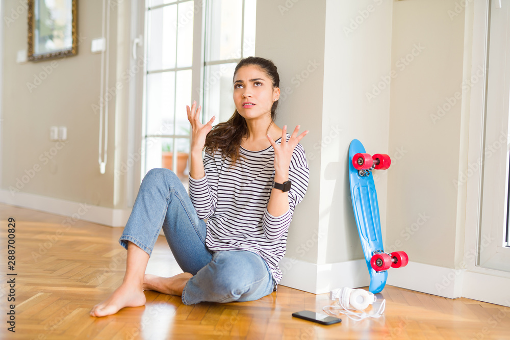 Poster young woman sitting on the floor using skateboard and headphones crazy and mad shouting and yelling 