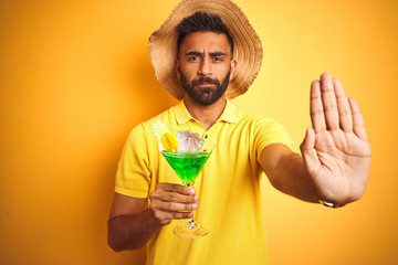 Indian man on vacation drinking cocktail wearing summer hat over isolated yellow background with...