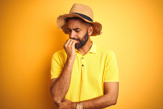 Young indian man on vacation wearing summer hat standing over isolated yellow background looking stressed and nervous with hands on mouth biting nails. Anxiety problem.