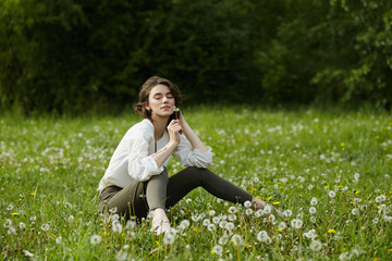 Portrait of a girl sitting in a field on the spring grass among dandelion flowers. Cheerful girl enjoys Sunny spring weather. Natural beauty of a woman, natural cosmetics