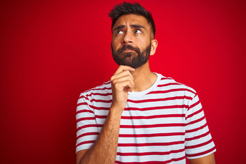 Young indian man wearing striped t-shirt standing over isolated red background with hand on chin thinking about question, pensive expression. Smiling with thoughtful face. Doubt concept.