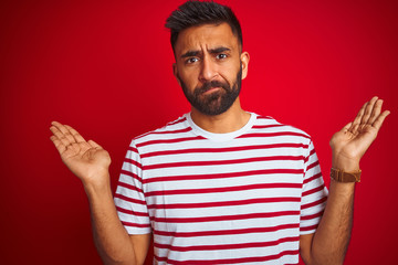 Fototapeta premium Young indian man wearing striped t-shirt standing over isolated red background clueless and confused expression with arms and hands raised. Doubt concept.