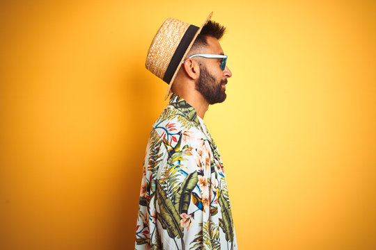 Indian man on vacation wearing floral shirt hat sunglasses over isolated yellow background looking to side, relax profile pose with natural face with confident smile.