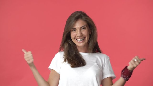 Extremely happy, cheerful adult woman with braces jumping from bottom, showing thumbs-up joyfully shaking hands, feeling upbeat and enthusiastic, triumphing from excellent news, red background