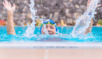 Head of an old lady peeks out of the pool water. Healthy pool activity. Partially hidden by the water spray. With cap and googles. People smiling and playing sports