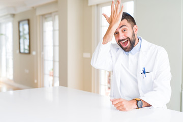 Handsome hispanic doctor or therapist man wearing medical coat at the clinic surprised with hand on...