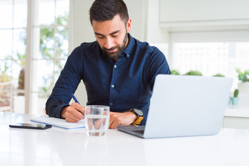 Handsome hispanic man working using computer and writing on a paper with a confident expression on...