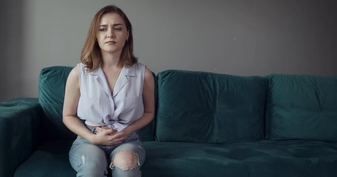 Young woman sits on the couch and feels belly pain.