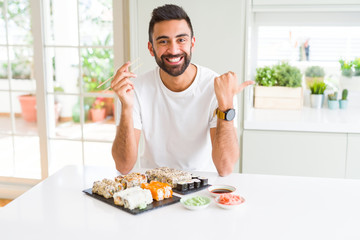 Handsome hispanic man eating asian sushi using chopsticks pointing and showing with thumb up to the side with happy face smiling