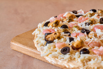 A part of raw homemade pizza with seafood and carbonara sauce on a wooden board. Cooking homemade pizza with shrimp, mussels, black olives and mozzarella. Closeup, selective focus